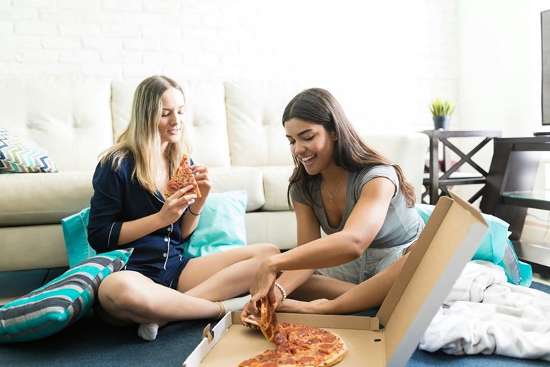 Two friends sit on the floor enjoying pizza together in a relaxed, cozy living room.
