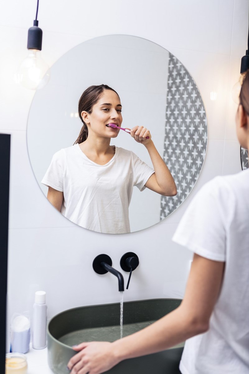Young beautiful dark-haired woman standing near sink while brushing her teeth