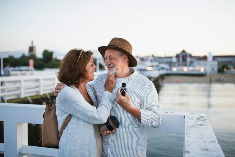 Older couple smiling at each other on a waterfront pier, with the woman touching the man’s face as he holds binoculars.