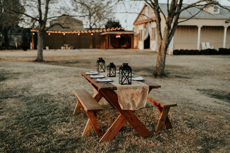 A rustic outdoor table set with lanterns and place settings sits in a quiet farmyard, with a softly lit barn and string lights glowing in the background.