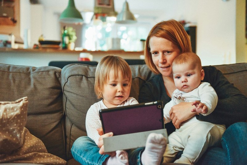 A mother sits on the couch with two young children, all focused on a tablet in a cozy living room.