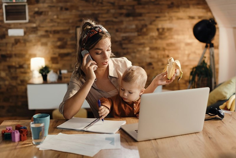 A mother juggles a phone call, work, and caring for her child at a desk, multitasking through a busy day.