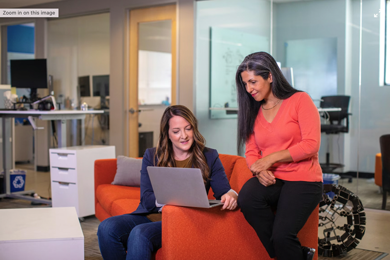 A wide shot of two female coworkers helping one another in the office.