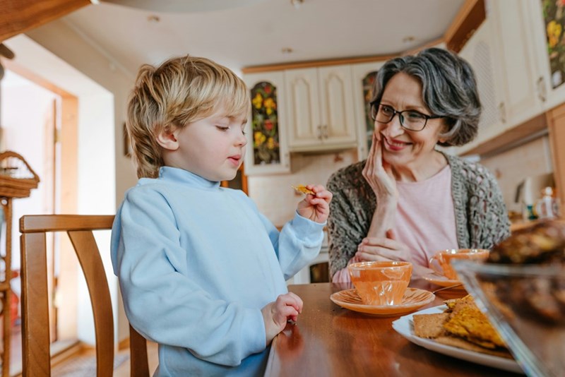 Grandma dotes on her 3-year-old toddler grandson as he eats a snack.