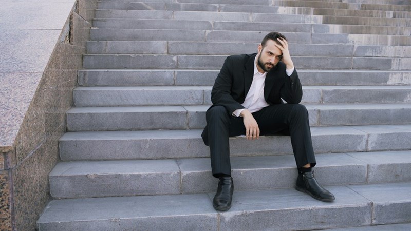 A frustrated employee puts his head in his hand as he sits on the front steps of his office building.