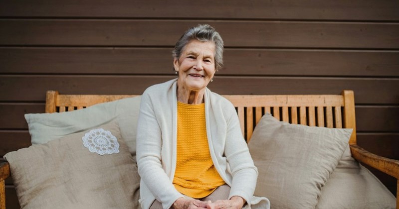 An elderly woman grins while sitting on her porch.