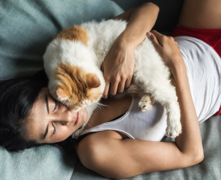 A cat is sleeping on a woman's chest and face, and she lovingly hugs the cat while they fall asleep together on a bed.