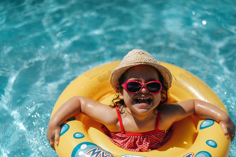 Little girl in red bathing suit, red sunglasses, and straw hat laughs with happiness while she floats in yellow pool floatie.