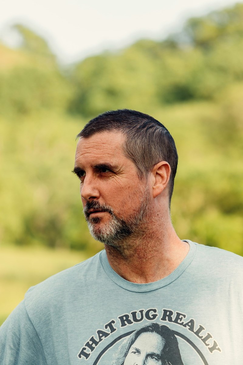 Middle aged an with short hair and a beard standing outdoors, looking off to the side with a blurred green background.