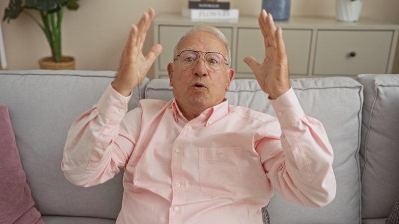 Elderly man sitting on a couch gesturing with raised hands, looking surprised or animated.