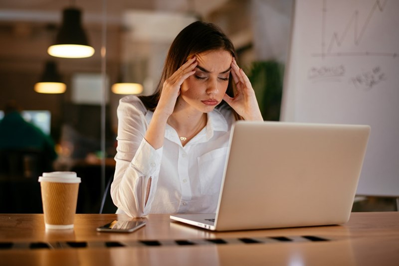 Woman sitting at a desk holding her temples while looking at a laptop, with a coffee cup and phone beside her.
