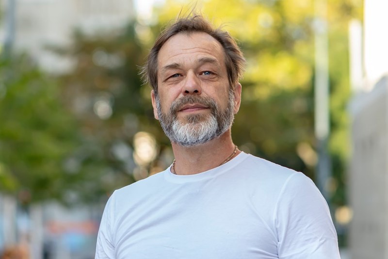 Middle-aged man with a short beard wearing a white shirt, standing outdoors and looking ahead.