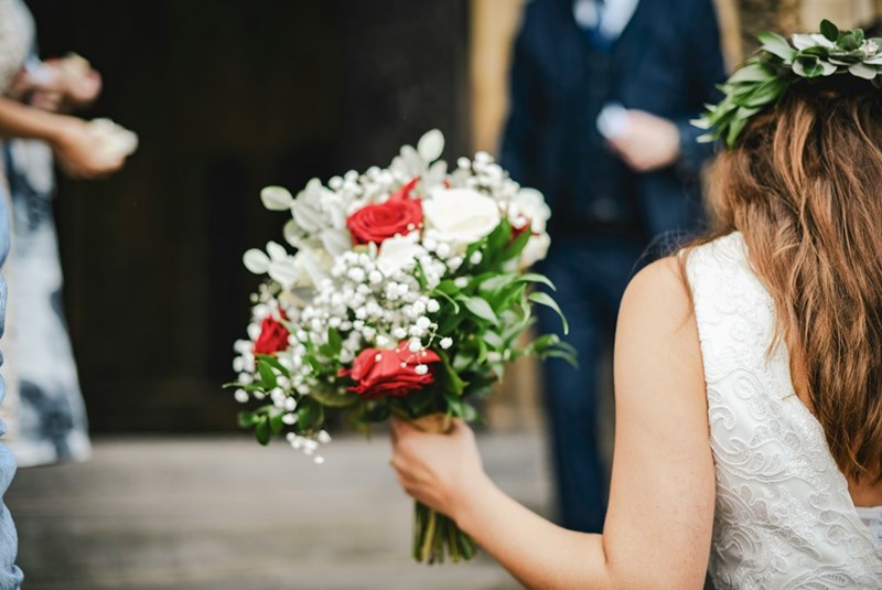 A bride holds a bouquet of red and white flowers while walking past blurred guests during a wedding moment.
