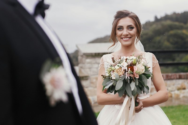 A smiling bride stands outdoors holding her bouquet, facing her partner during an intimate ceremony scene.