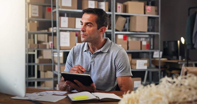 Man in a warehouse holding a tablet and looking to the side, with shelves of boxes in the background.