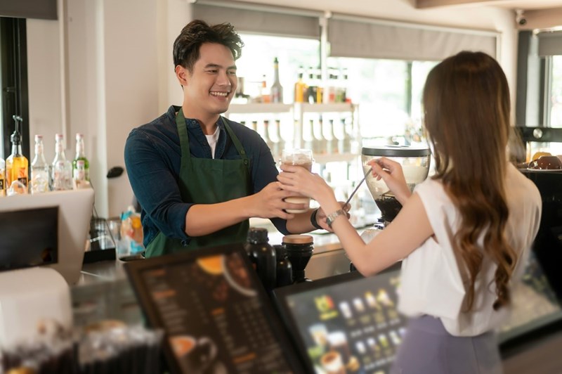 Young male barista serves customer a glass of iced coffee