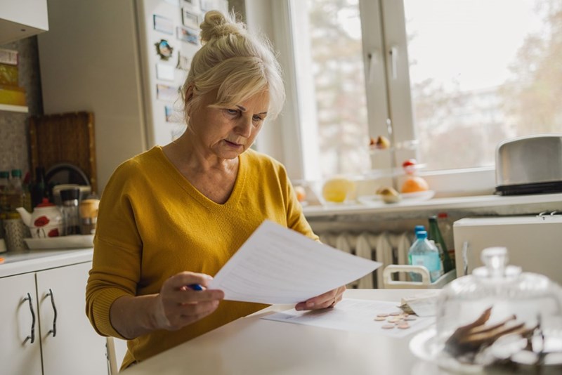 Older woman sitting at a kitchen table reviewing a document, with coins and household items nearby.