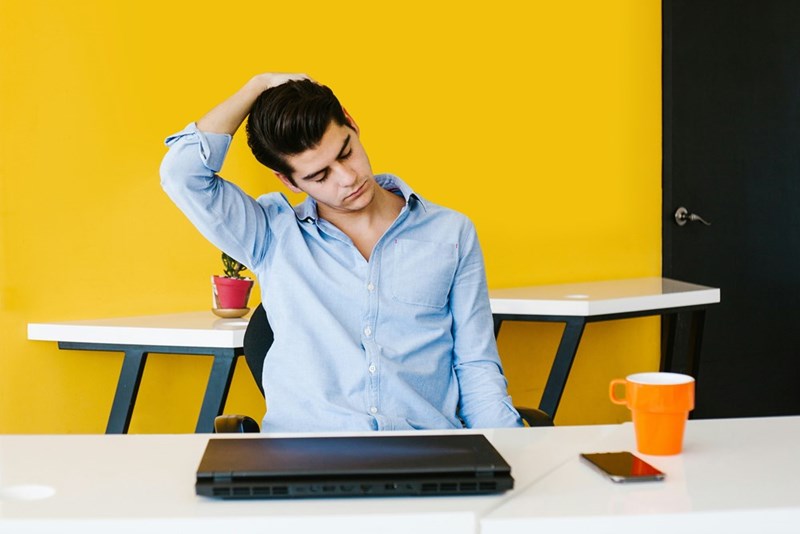 Office worker holds his neck while a closed laptop sits in front of him, as he contemplates his mistakes.