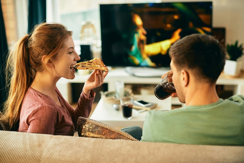 Teen couple eats pizza and drinks soda while sitting on living room couch watching TV.