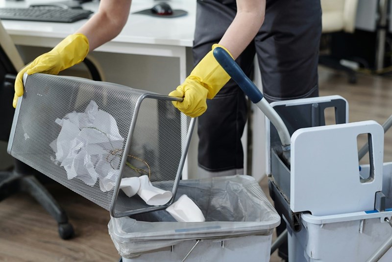 A cleaner wearing gloves discards trash into a garbage bin in an office.