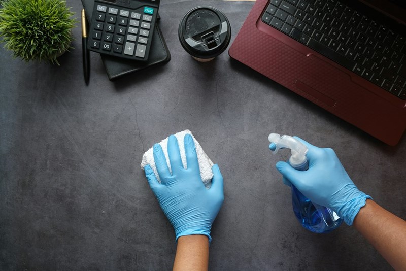 A cleaner wearing gloves wipes an employee's desk in an office.