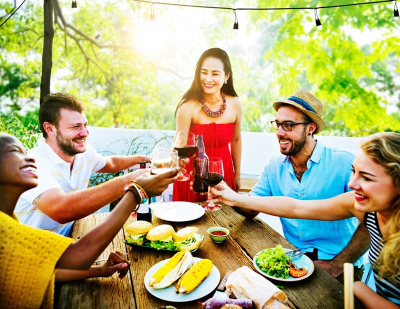 A group of friends laugh and toast together during a sunny outdoor meal.