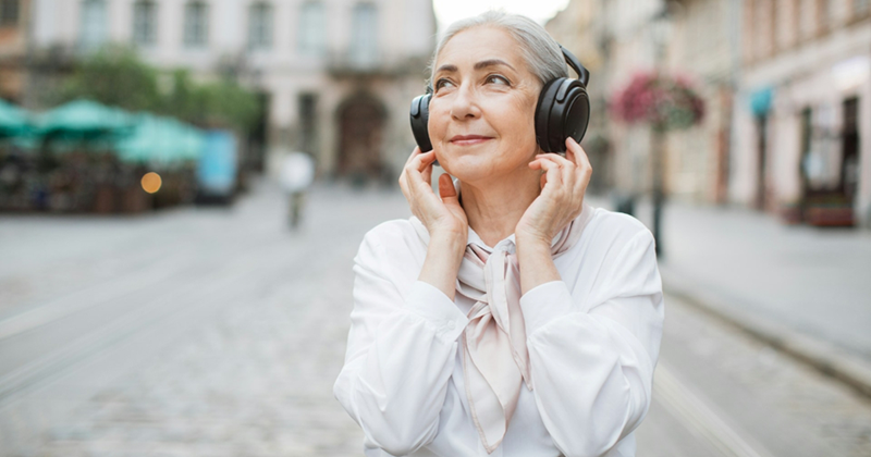 An older woman smiles slightly while wearing headphones and standing in the middle of a street