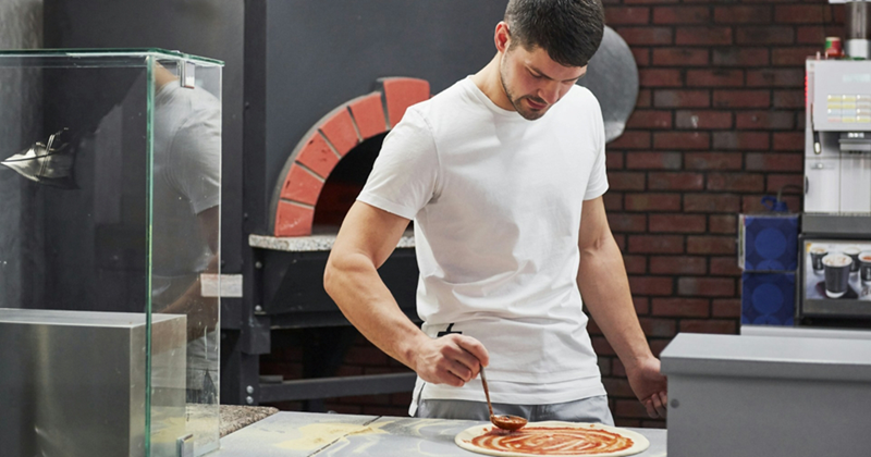 A man puts sauce on a pizza base with a wood-fired oven behind him