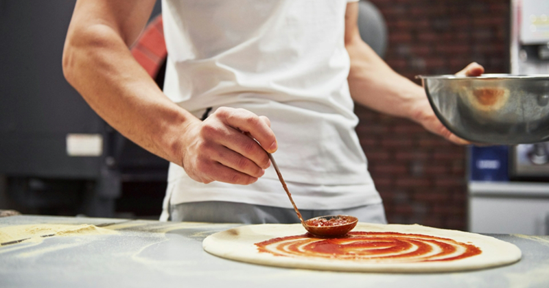 Close-up of a man holding a metal bowl and using a ladle to put sauce on a pizza base