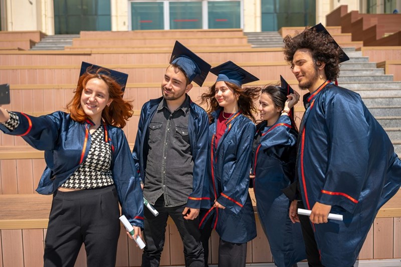 Group of university graduates in blue gowns taking a selfie after graduation on campus steps