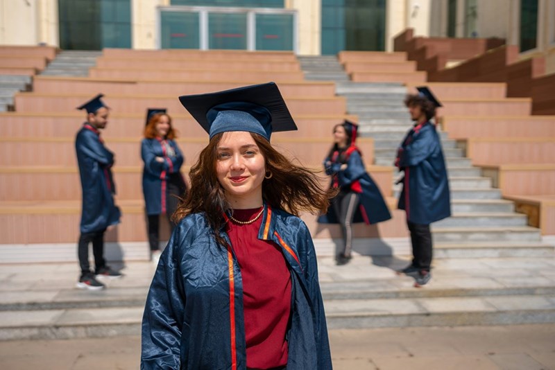 Confident female college graduate posing in cap and gown with classmates in the background on graduation day