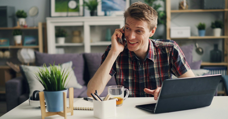 A man sits at a desk with his laptop in a home office, smiling as he takes a call on his cellphone