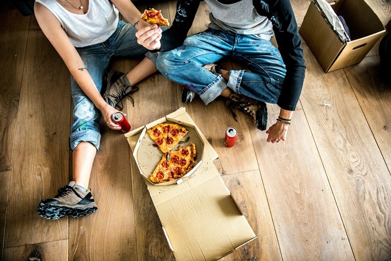 Two young roommates sitting on the floor sharing pizza and drinks in a casual apartment setting