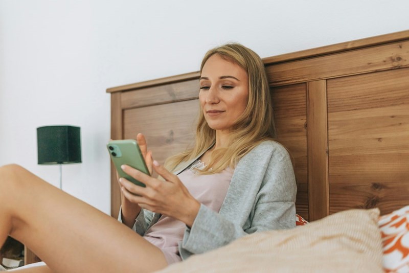 Woman sitting on a bed, relaxed, looking at her phone with a slight smile.