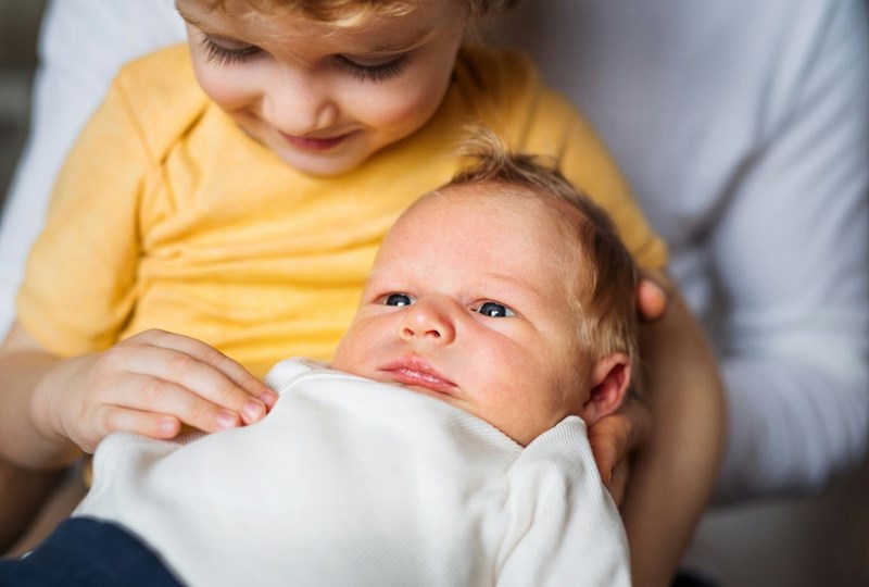 A small toddler boy holding a newborn baby brother at home