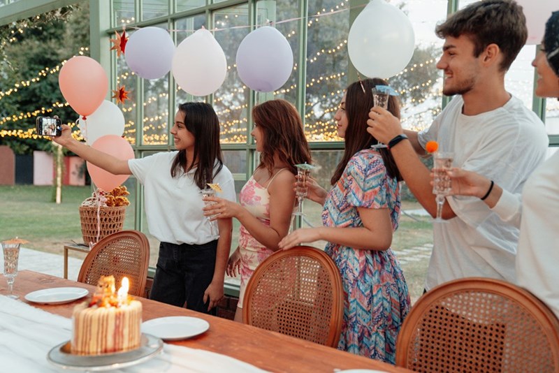 Guest takes a selfie with other guests and the birthday girl at her lavish event, with a cake with lit candles is on the table in front of them.