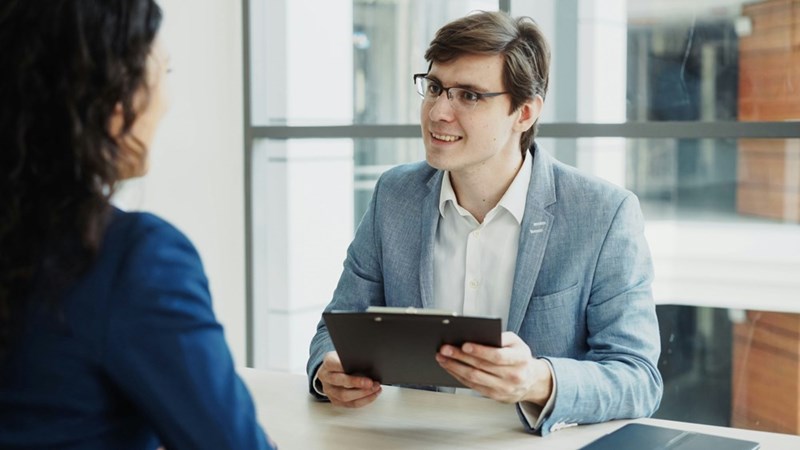 A man in a suit holds a clipboard while interviewing a job candidate, smiling awkwardly.