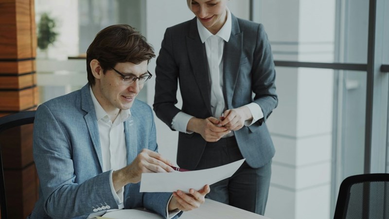 A man in a suit points to a document as a woman stands next to him, looking over his shoulder.