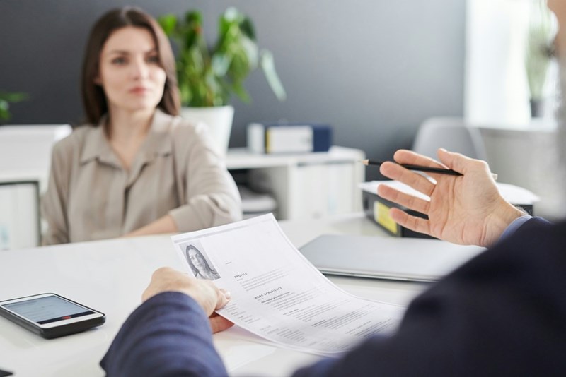 A manager evaluates his female employee's paperwork as she sits across from his desk.