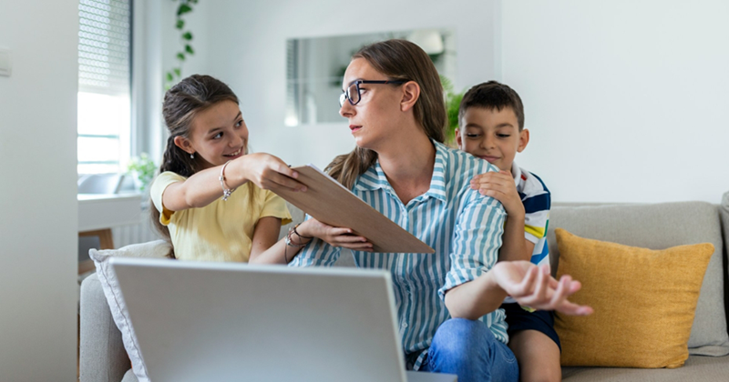 A mom working on a laptop while sitting on a couch shrugs as her daughter hands her a clipboard, her son clinging to her other shoulder