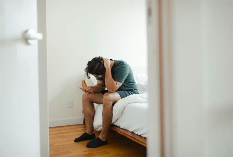 Regretful man sits on the side of a bed sending phone messages.