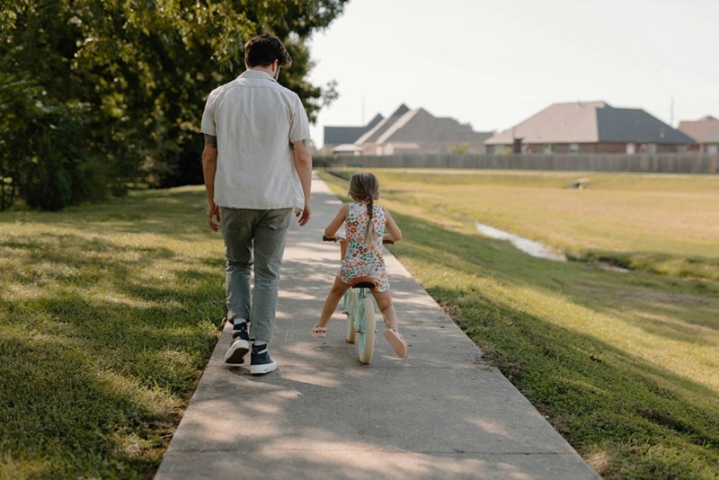 Dad walks alongside daughter as she rides her bike down the sidewalk.