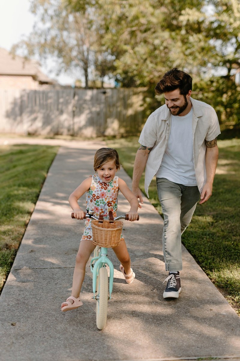 Little girl rides her bike down the sidewalk while Dad runs alongside her.