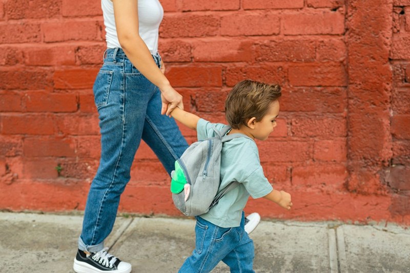 Little boy wearing backpack holds Mom's hand as they walk to school.