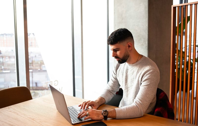 Man sits at a wooden table by large windows, typing on a laptop with focused attention in a bright, modern workspace.