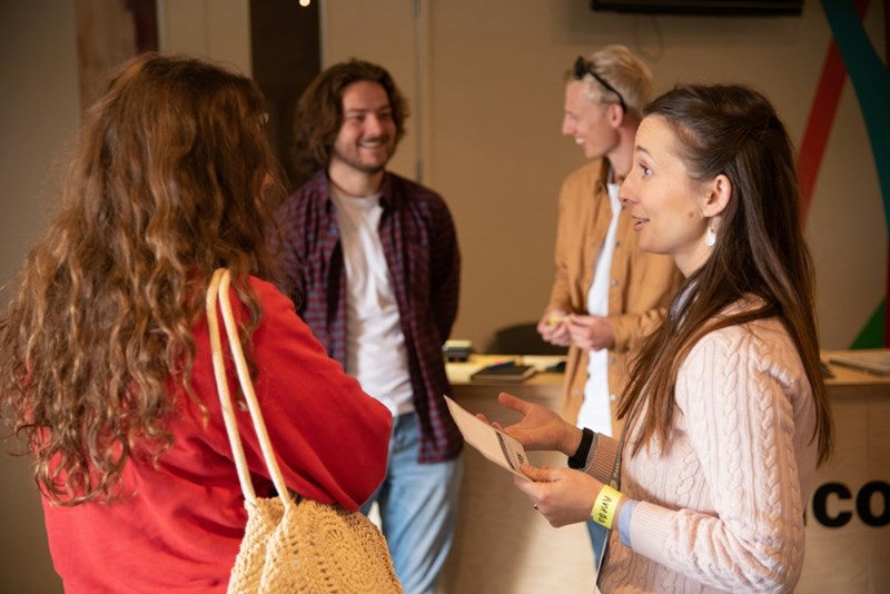 Group of adults chatting indoors, one woman holding paper during casual networking conversation.