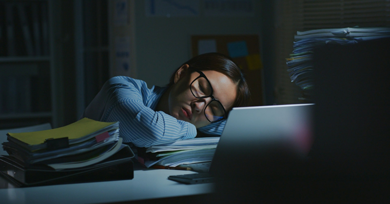 A young woman wearing glasses sleeps on a desk surrounded by piles of papers