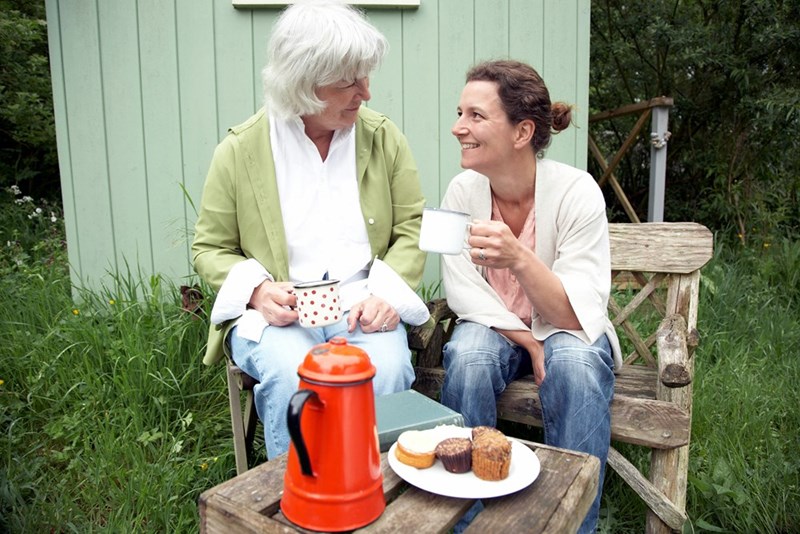 two women sitting in front of a shed on a wooden bench outdoors, smiling and chatting while holding mugs beside a small table with pastries.