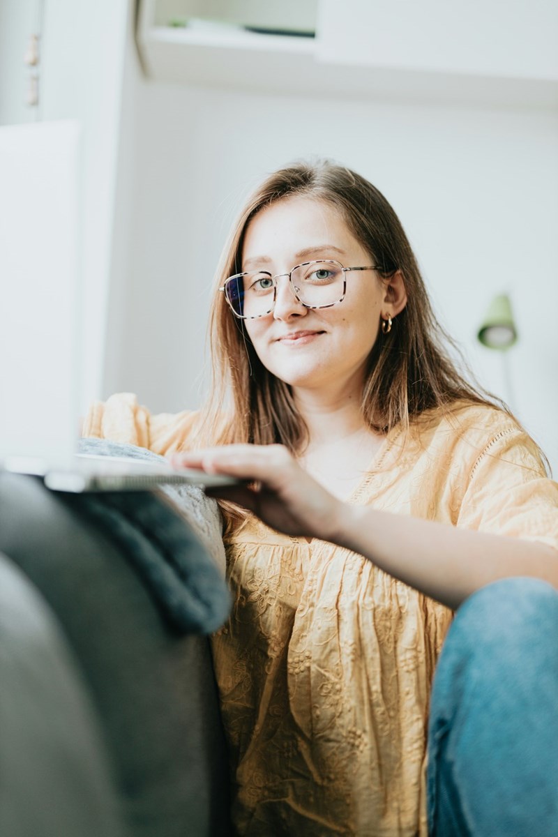 Woman wearing glasses sits on a couch, smiling while using a laptop in a bright living room.