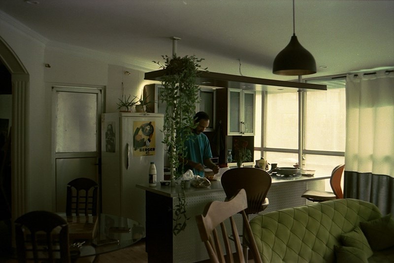 Man stands in a dimly lit kitchen preparing food at a counter, surrounded by plants and warm indoor lighting.
